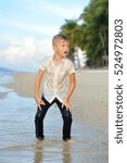 Small photo of Full growth portrait on a tropical beach: handsome 8 years old boy in wet clothes without shoos ankle-deep in water. Leaned his hands on legs, ready for a jump, looking into the distance