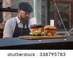 Small photo of KIEV, UKRAINE - 16 AUG 2015: Festival street food. Two hamburger stand on the board with tags. In the background, cook with a beard, wearing glasses and a cap.