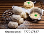 Small photo of Six lemon crinkle cookies stacked in the form of a pyramid and some Aunt Birdie's cookies with a green cherry. It is displayed with a sieve, whisk and castor sugar on a striped wooden board